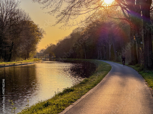 Fotografie Cyclist on canal towpath at golden hour sunset in Rijkevorsel, België - peaceful
