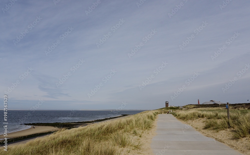 Fototapeta premium Wide-angle view of a coastal pedestrian path leading toward a lighthouse and observation tower on the sand dunes of Den Helder, Netherlands.
