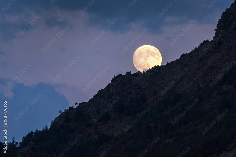 Fototapeta premium Full supermoon rising behind mountain slope, large bright moon over landscape in clear night sky