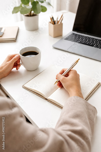 Minimalist home office workspace with hands writing in notebook, holding coffee mug, laptop on desk, soft natural light, clean and modern productivity concept.