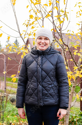 Portrait of a smiling middle-aged woman wearing a warm black puffer jacket and a knitted hat. Standing outdoors in an autumn garden with yellow leaves background