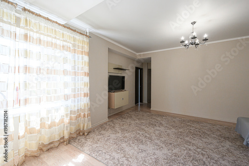 A light-filled living room with beige curtains, a patterned carpet, a TV unit, a chandelier, and light wooden flooring. Neutral tones, spacious, airy, and well-lit