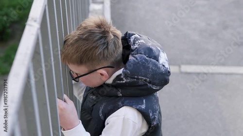 A teenage boy stands behind a metal fence, looking upset and isolated. The scene conveys sadness and bullying at school, highlighting emotional struggle and vulnerability. 