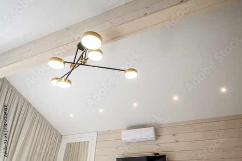 A modern ceiling chandelier with gold-rimmed circular lamps and black metal arms hangs in a light-toned room with wooden paneling and recessed ceiling lights