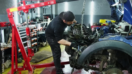 Young mechanic dismantles car engine with hydraulic hoist in repair shop slow motion. Man technician lifts motor with automated equipment in workshop