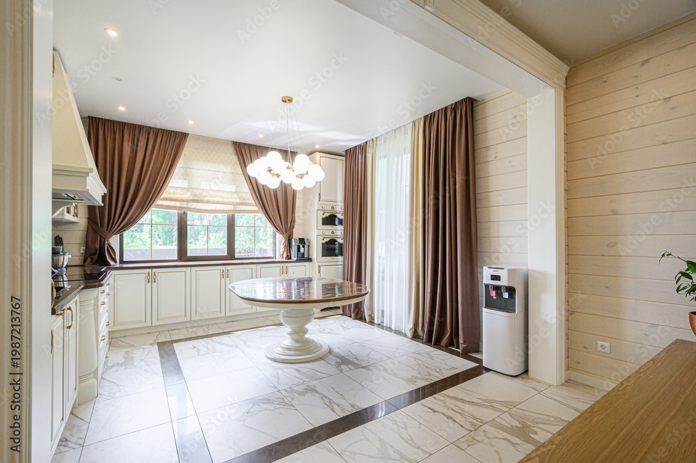 Fototapeta premium Elegant kitchen-dining area with marble flooring, white cabinetry, round dining table, chandelier, large windows with brown curtains, light wood walls, and water dispenser, bright and cozy.