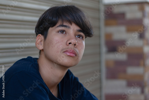 A teenage Asian boy sitting alone indoors, resting his arms on his knees with a sad and contemplative expression. Soft natural light and shallow depth of field emphasize his face and realistic skin te