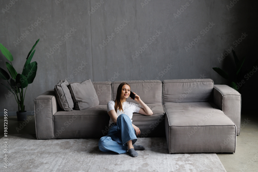 Obraz premium Young pretty woman talking by mobile phone on carpet near grey sofa at home