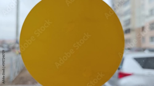 Yellow circular object gradually shifts in position against a blurred urban backdrop, revealing details of its surface and surrounding environment in three sequential frames