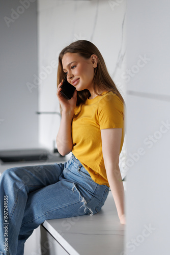 Young woman using smart phone while sitting at the kitchen counter