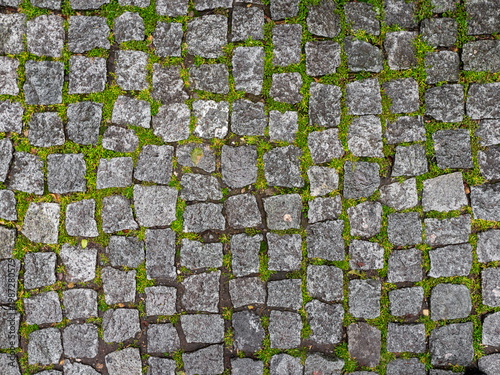 Old cobblestone pavement with green moss and grass. Top view of granite stone road texture background