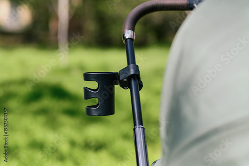 Close-up of stroller handle with black cup holder attachment in sunny park, shallow depth of field with green background, modern parenting accessory and outdoor lifestyle concept for advertising use
