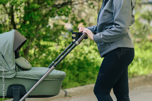 Close side view of woman pushing baby stroller with cup holder in green park, focus on hands and handlebar, calm parenting routine and modern childcare lifestyle concept for advertising