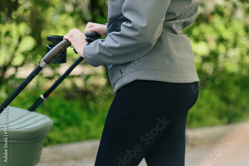 Cropped rear side view of woman pushing baby stroller in green park, focus on hands on handle with cup holder, soft natural light, everyday parenting routine and modern lifestyle concept