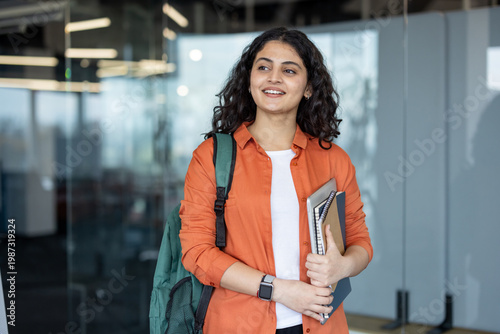 Young indian woman carrying a backpack, laptop, and notebooks, smiling and looking away while walking through a bright modern university campus building