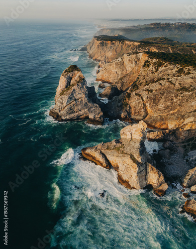 Aerial view of dramatic rocky coastline with cliffs and turquoise ocean waves crashing against rugged shore at golden hour. Portugal