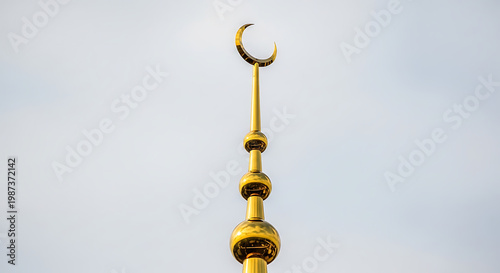 Tall golden minaret spire topped with islamic crescent moon symbol against clear pale sky, architecture of mosque, religious landmark, high quality photography with sharp details and realistic light.