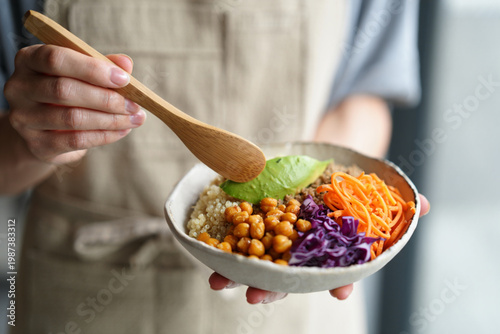 Close-up of person in linen apron assembling healthy Buddha bowl with avocado and wooden tongs, vibrant organic ingredients in kitchen