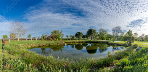Charencey, France - 04 10 2026: Panoramic view of a pond, large trees, grass and a blue cloudy and sunny sky in a park in Le Perche region