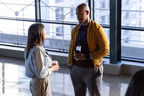 Diverse coworkers chatting in atrium by floor-to-ceiling windows holding coffee cups with lanyard