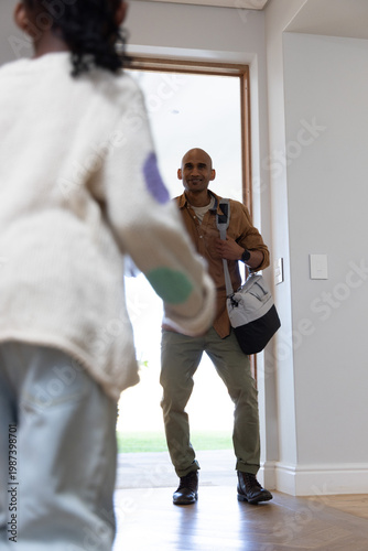 Diverse dad and child greeting in home entry, dad carrying duffel, child wearing patched sweater