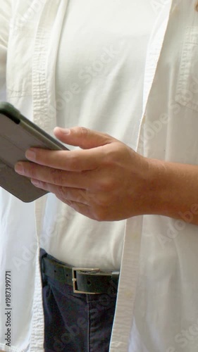 Vertical video: Framing adult in white tee adjusting, tapping folio tablet by window while reading