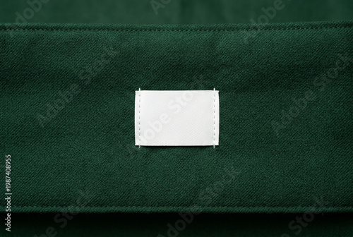 Small white blank rectangular tag sewn onto dark green textured cloth fabric surface mockup realistic.