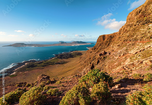 Sunlit volcanic cliffs in Lanzarote overlook deep blue ocean, rugged shoreline, and distant islands beneath a clear sky in a dramatic coastal landscape.