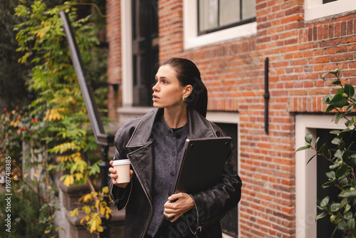 Thoughtful young brunette woman in black leather jacket walking on city street holding laptop and paper coffee cup, serious female freelancer looking away with focused expression outdoors near house