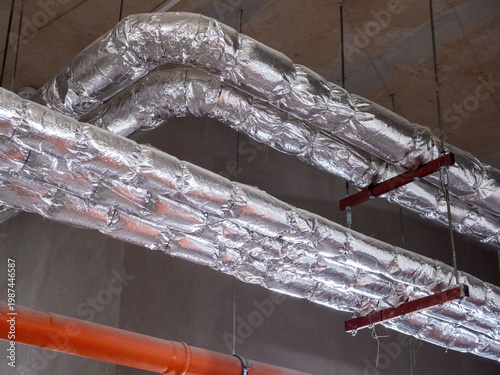 Industrial air ducts with silver thermal insulation, orange pipe on ceiling. Large air ventilation ducts are wrapped in shiny reflective silver thermal insulation foil, suspended from concrete ceiling