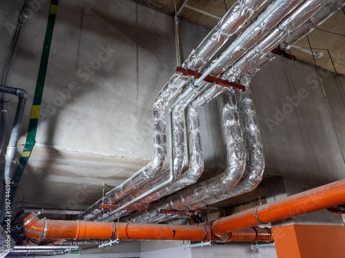 Industrial utility systems with silver insulated ducts and orange drainage pipes on ceiling