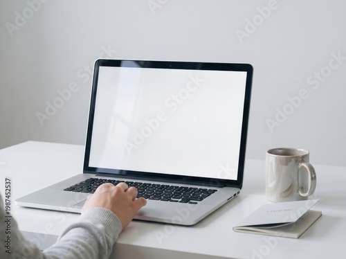Person using a laptop computer with a blank screen on a white desk with a notebook and coffee mug