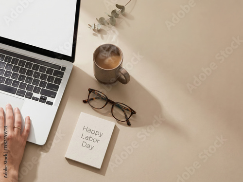 A persons hand on a laptop keyboard with a happy labor day note, coffee, and glasses on a beige desk