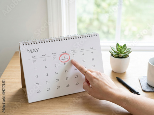 Hand pointing to may 1st on a desk calendar with a red circle around the date, sitting on a wooden desk with a plant, coffee cup, and pen nearby