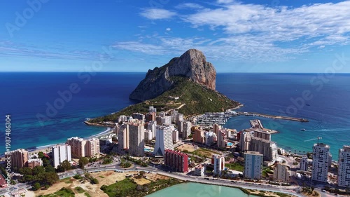 Aerial panoramic view of Calpe, Spain, featuring the iconic Penyal de Ifac rock formation rising above the coastal city, turquoise Mediterranean waters, marina and salt lagoon