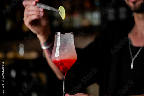 A bartender uses tweezers to place a lime slice garnish on a red and pink ombre cocktail in a chilled stemmed glass.