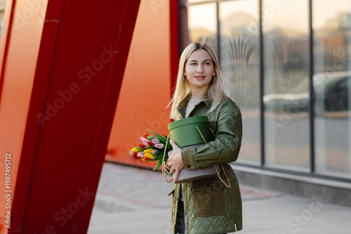 Canvas Print Young woman carrying tulips and green hatbox walking near red modern architectur