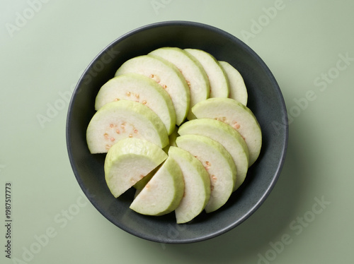 Top view of fresh sliced white guava in a dark bowl on a pale green background