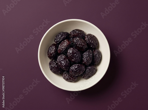 Top view of whole dried prunes in a light bowl on a dark purple background