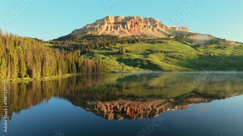 Stunning mountain peak with red rock cliffs reflects perfectly in glass-like alpine lake water. Morning mist rises over evergreen forest as golden sunrise light illuminates the rugged landscape.