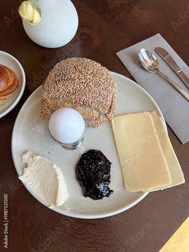 Healthy breakfast plate with boiled egg, cheese, and jam.

 A nutritious morning meal featuring a soft-boiled egg, fresh brown bread roll, a slice of cheese, cream, and dark fruit jam on a