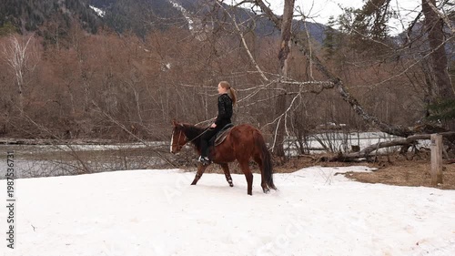 A girl rides a horse in the snow against the backdrop of a mountain river and snow-capped mountains.