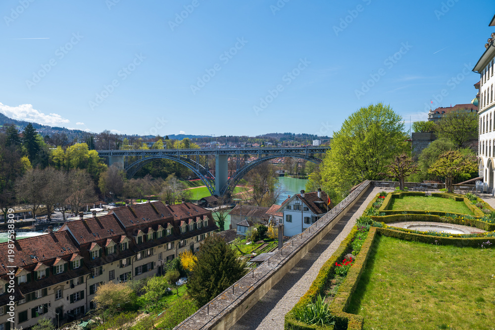 Obraz premium Panoramic spring cityscape of Bern with the Aare River, Kirchenfeld Bridge, old town rooftops and terraced gardens under a clear blue sky.