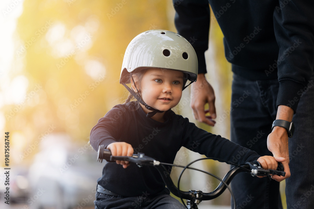 Obraz premium Father guides young caucasian daughter learning to ride bicycle outdoors