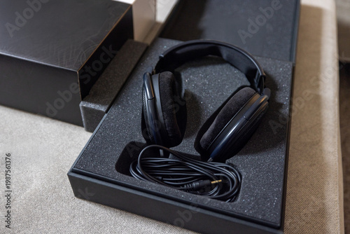 Black over-ear headphones sit in open packaging. Cable lies neatly coiled beside them. Box rests on textured beige surface. Clean, minimalist product shot