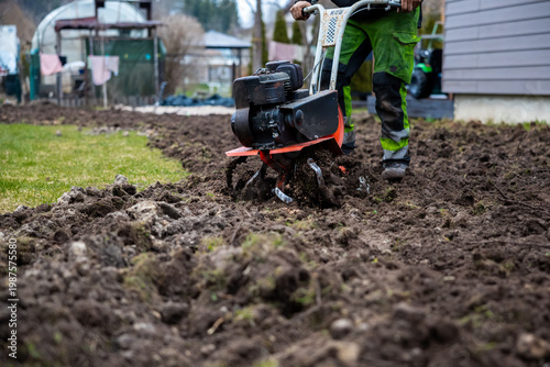 A person operates a red and black rotary tiller, churning wet soil into deep furrows beside a neat lawn near a residential structure during early spring.