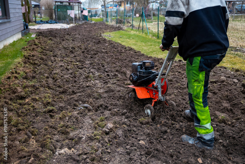 Gardener in high visibility green pants tills compacted soil by a house and fenced yards. Overcast light and damp ground suggest cool pre planting work and rough soil texture.