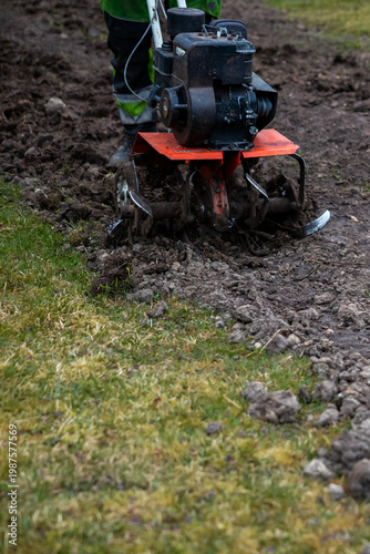 A groundskeeper in high visibility pants steers an orange deck tiller, tines churning damp soil beside mown grass in a garden plot during cool weather with muted light.