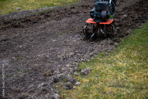 Obraz na plátně A person runs a compact rotary tiller with a red deck and exposed tines, cutting