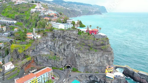 Aerial view of Ponta do Sol, Madeira. Colorful buildings cling to cliffs overlooking the Atlantic, with a charming fishing village and dramatic coastline under a cloudy sky.